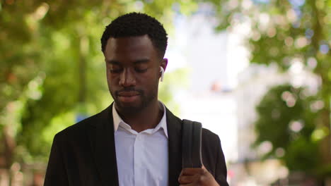 young businessman wearing wireless earbuds streaming music or podcast checking mobile phone walking to work in offices in the financial district of the city of london uk