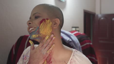 happy and hopeful cancer patient bald girl applies colored powder on cheeks at home on holi, indian festival of colors, slow motion