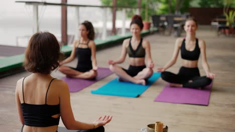 Group-of-girls-sitting-on-the-outdoors-wooden-floor-in-lotus-pose,-yoga-led-by-trainer