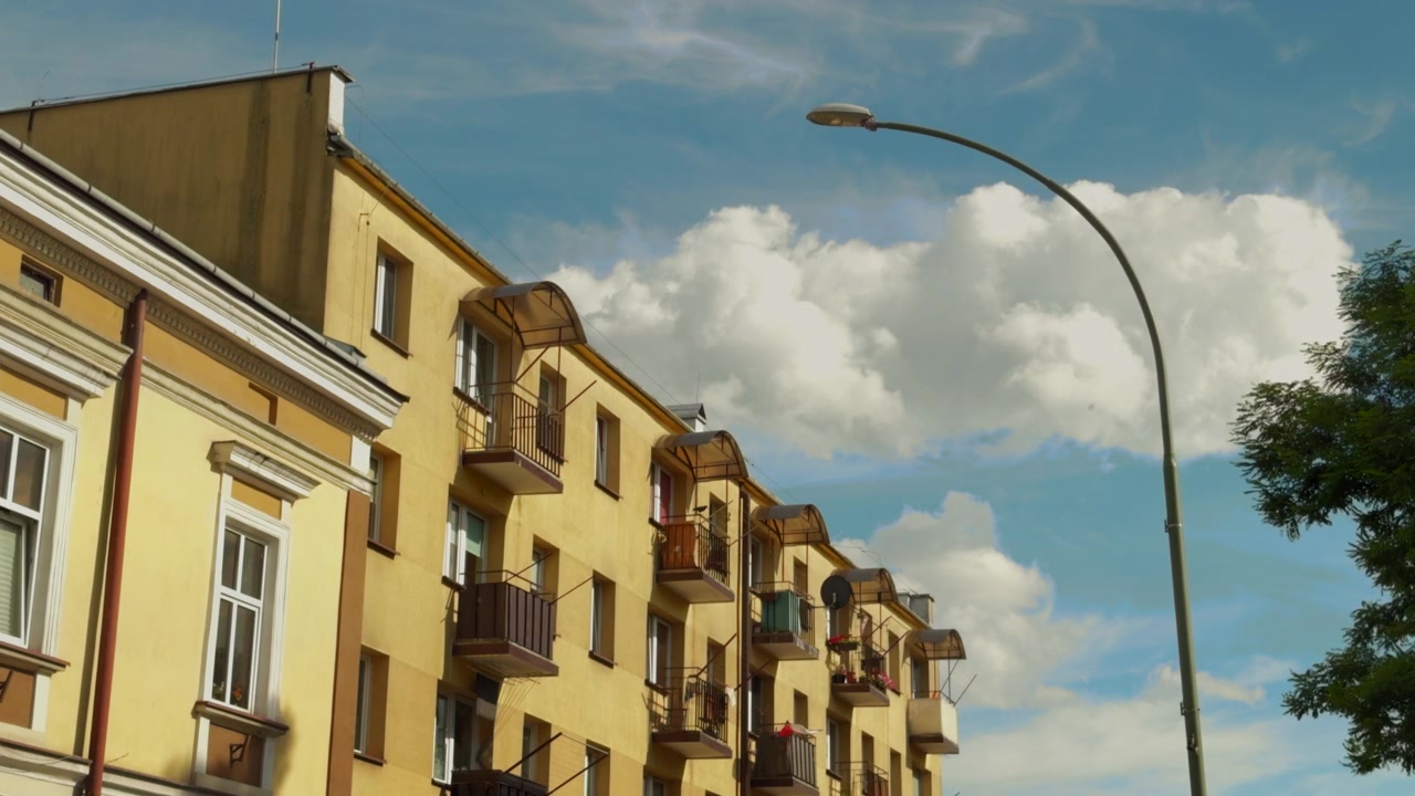 Toma estática de ángulo bajo de edificios de apartamentos residenciales amarillos de Europa del Este con balcones en Sanok, Polonia, enmarcada por una farola curva bajo un cielo azul con nubes cúmulos dramáticas.