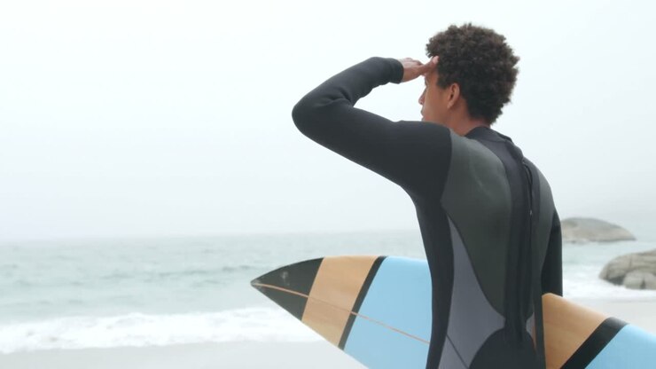 Vista posteriore di surfista afroamericano in piedi con la tavola da surf sulla spiaggia 4k