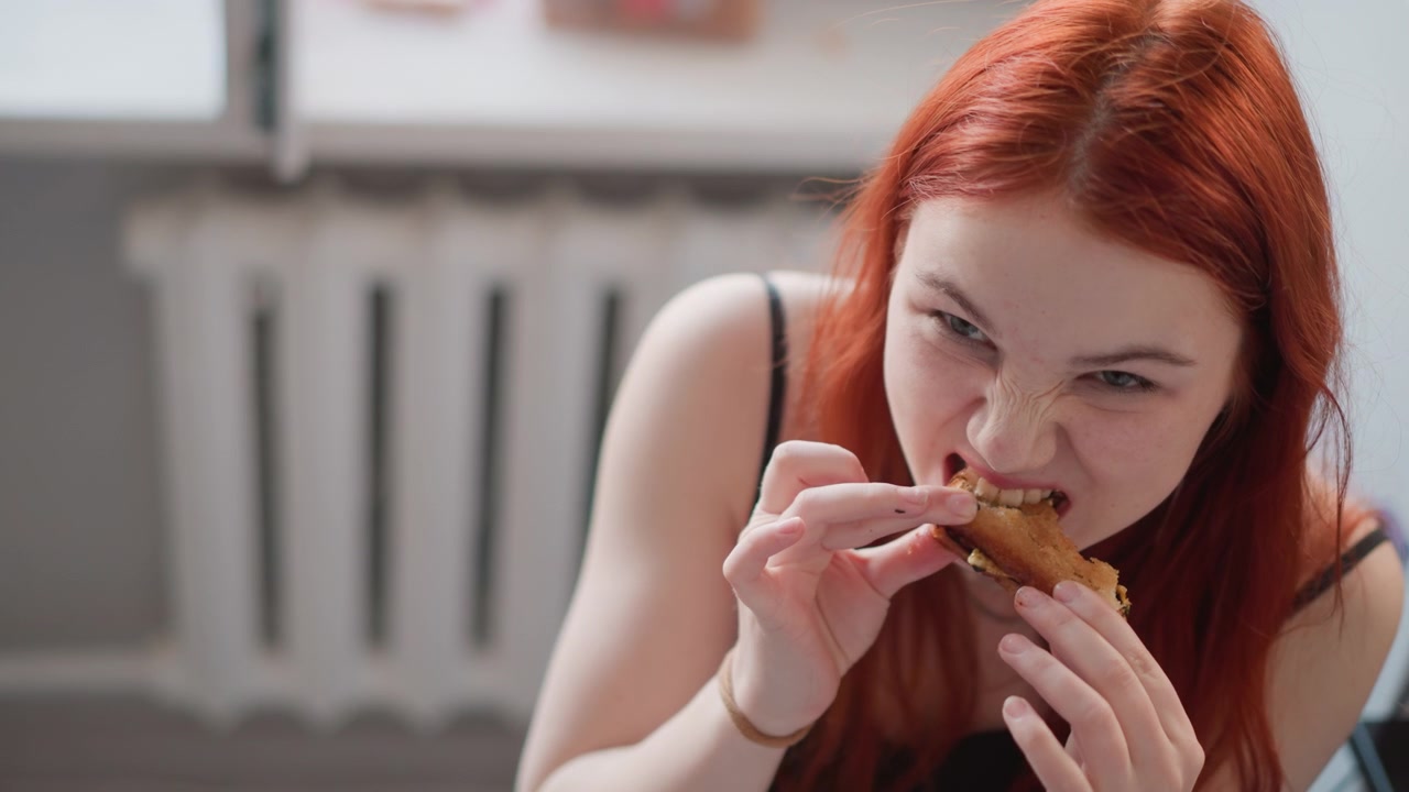 Young woman eating sandwich with gusto, energetic messy devour shows expressive face, crumbs and filling falling, casual apartment setting with radiator background, high energy bite conveying hunger