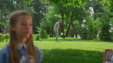 young father passing ball active son behind resting daughter in park.