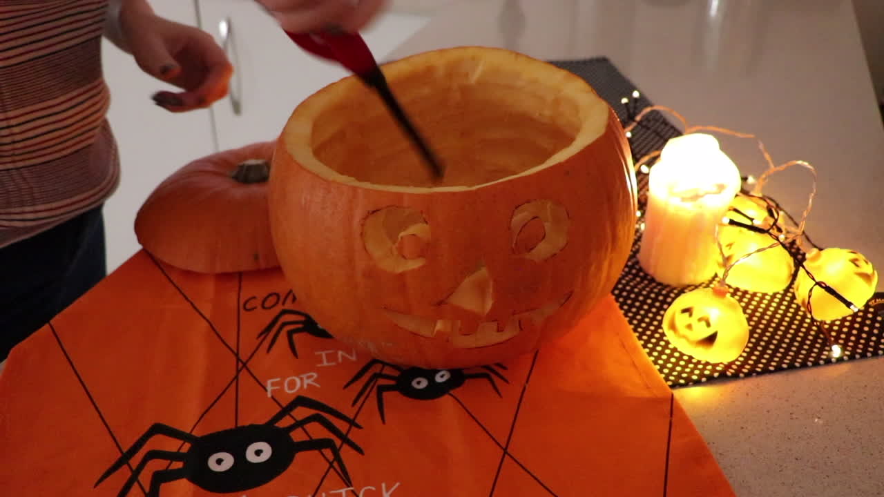 Woman lighting a hand carved orange pumpkin lantern at home in kitchen. With halloween lights and halloween themed table cloths
