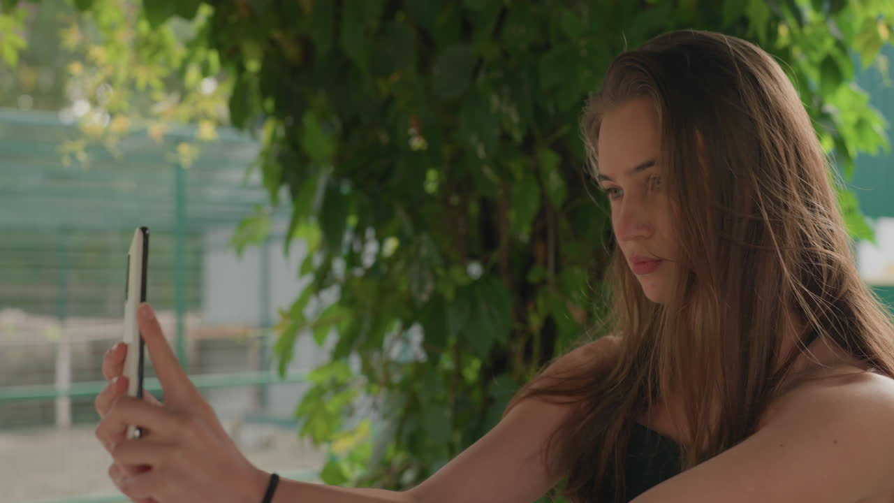 adolescente segurando um smartphone sob uma trepadeira, luz solar suave e plano de fundo verde, ajustando a câmera frontal com olhar concentrado, vestido de verão casual, preparando um retrato, expressão serena e natural