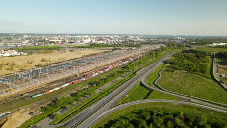 Luftaufnahme-Eines-Güterzugs-Bewegt-Sich-Auf-Der-Eisenbahn-Vom-Frachtterminal-Danzig-Aus,-Autoverkehr-Auf-Der-Schnellstraße-Im-Sommer,-Stadtpanorama-Danzig