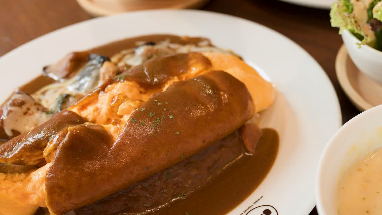 Hand slices fluffy omelette rice topped with creamy sauce, brown gravy, soup, and salad