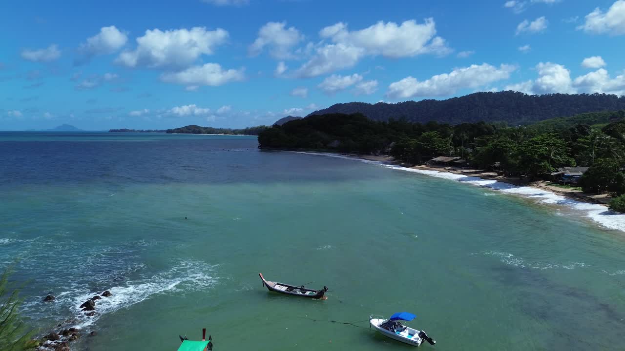 Cinematic aerial drone flight above dense tropical forest in Koh Lanta, Thailand, showing colorful longtail boats anchored in emerald water and revealing paradise beach with palm trees and bungalows