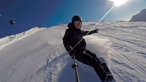 vista frontal de un snowboarder bajando a gran velocidad con un palo de selfie y teniendo una gran ola de nieve en la cara en un día despejado