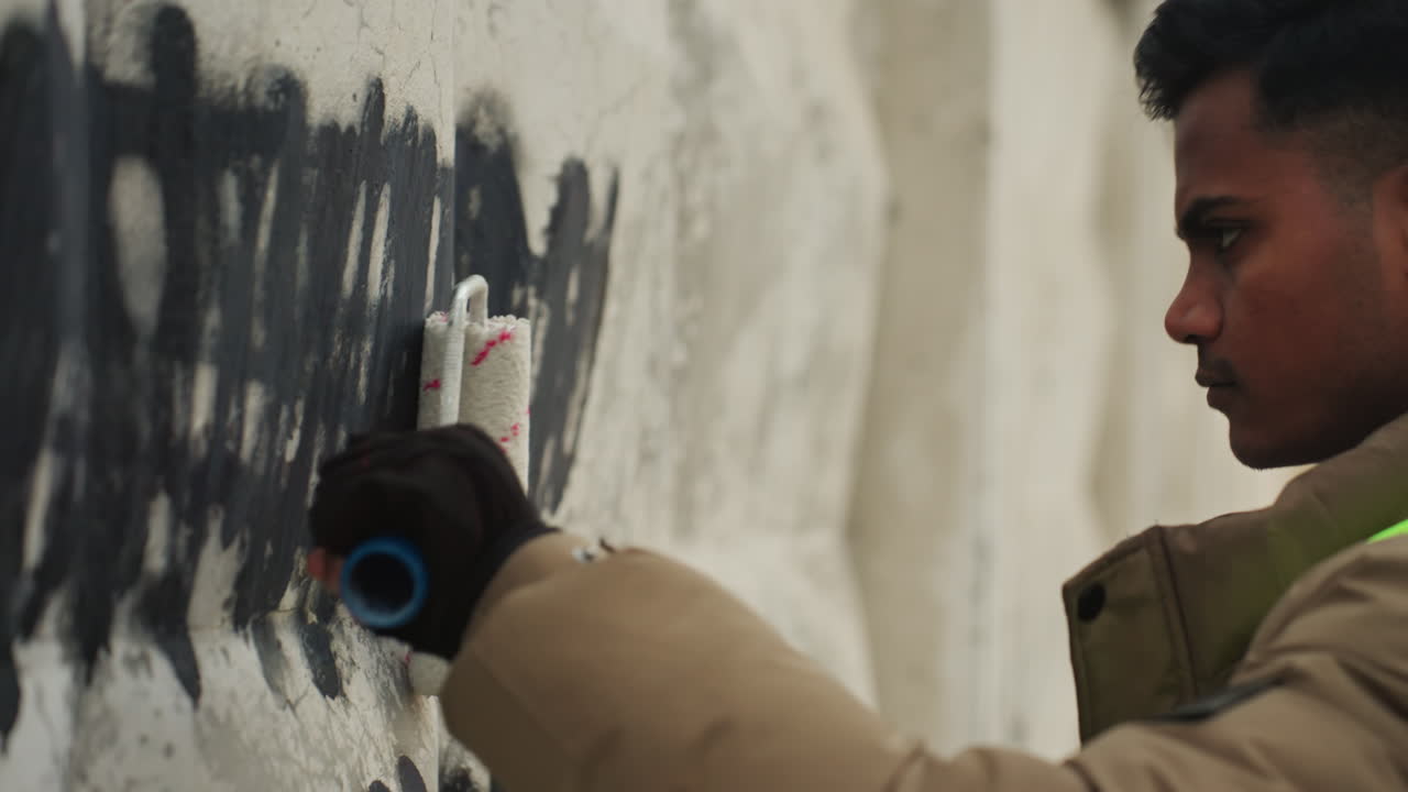 Sydasiatisk mand fjerner graffiti på væg. En byvedligeholdelsesarbejder iført refleksvest og tyk frakke bruger spraydåse og handsker til at dække sorte tags med et fokuseret udtryk i det kølige vinterlys.