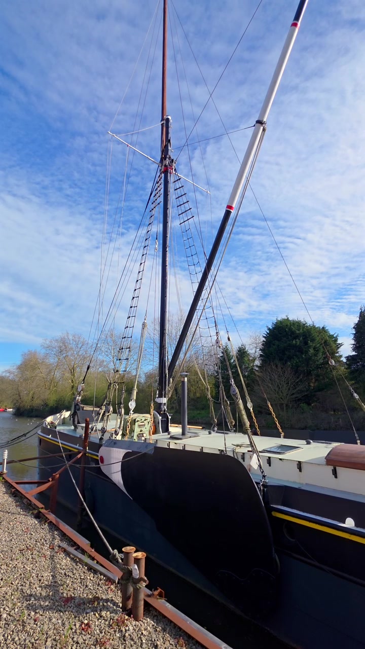Inquadratura statica di una barca a vela classica ormeggiata su un fiume calmo sotto un cielo azzurro.