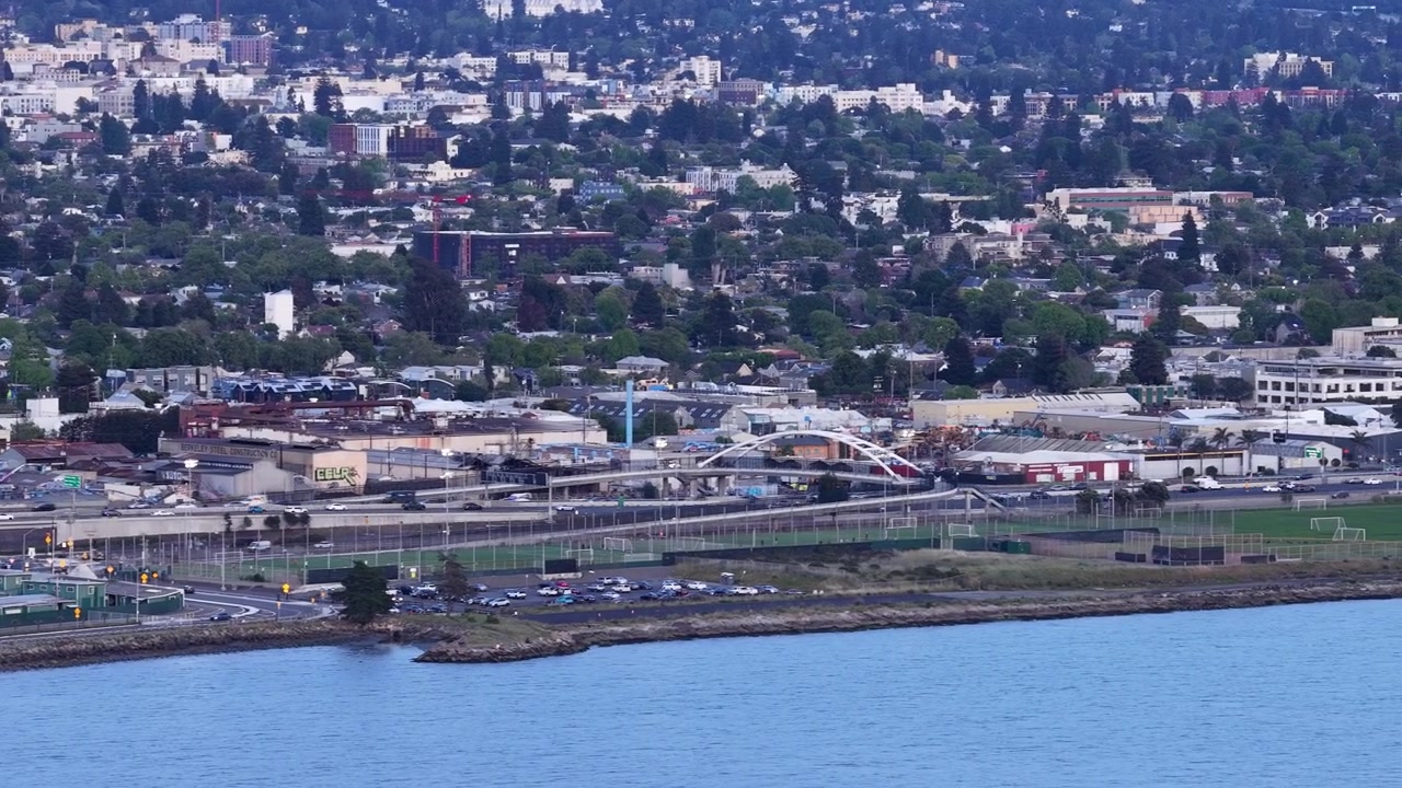 Ampia prospettiva aerea che mostra il paesaggio urbano di Albany e Berkeley, California, con le acque della baia in primo piano e il traffico che si muove lungo l'infrastruttura autostradale costiera