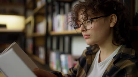 Close-up-an-interested-girl-with-curly-hair-and-glasses-sits-near-shelves-with-books-and-reads-one-in-the-library