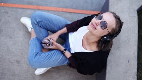 Attractive-girl-in-sunglasses-and-headphones-sitting-on-the-concrete-surface-with-crossed-legs.-Footage-from-the-top.-Girl