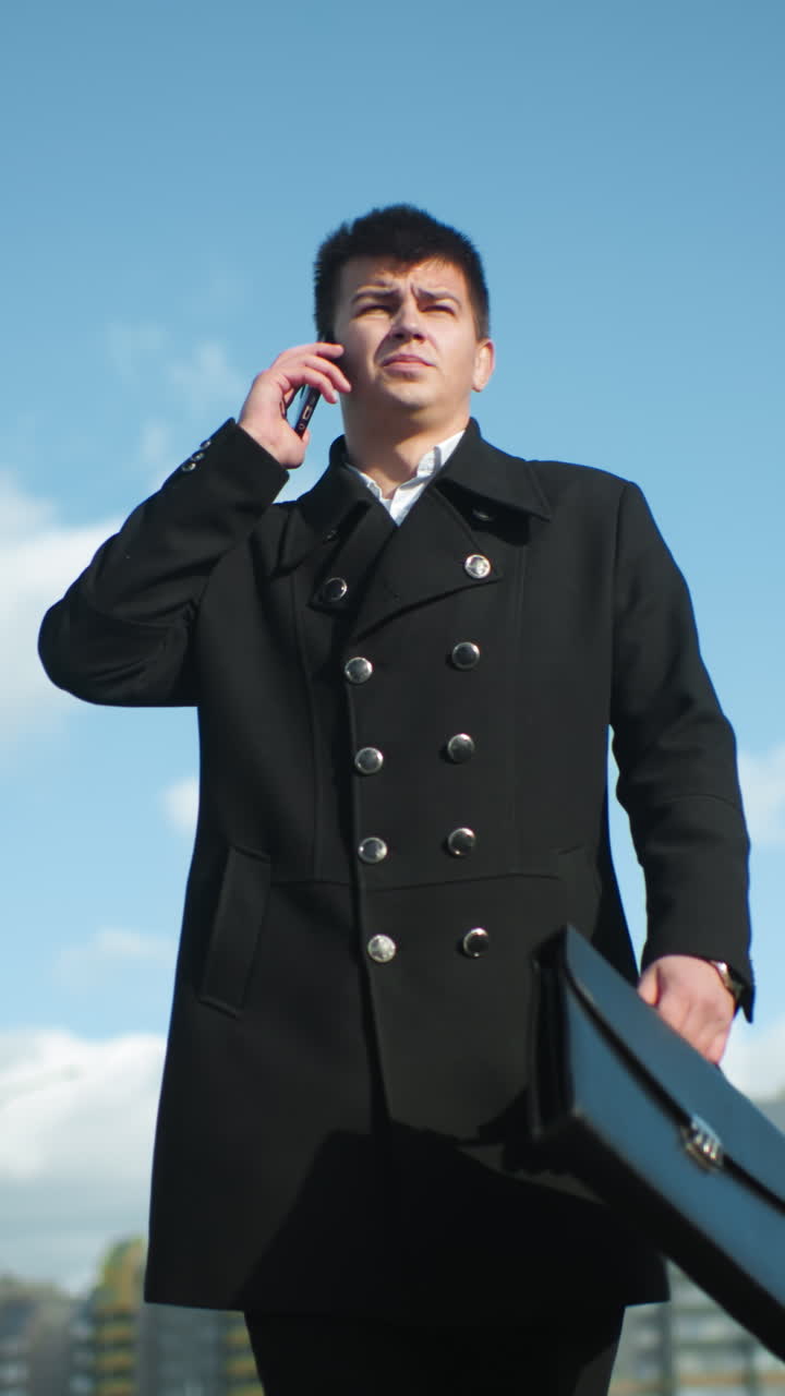 검은 코트를 입은 은행가가 주머니에서 전화를 꺼내서 전화에 응답하는 동안 현대적인 건물과 초록색이 흐릿한 배경에서 보이는 도시 환경에서 외부에서 자신감 있게 고 있습니다.