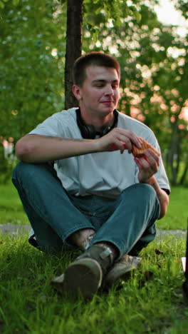 two students sitting on grass outdoors with laptop in front, toasting with bread in hand, surrounded by trees with sunlight filtering through them