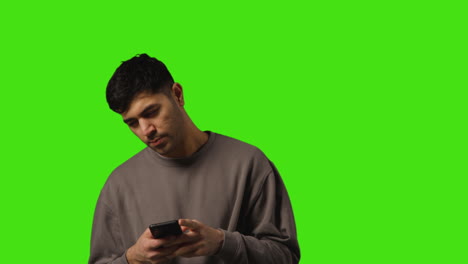 Young-Man-Writing-And-Sending-Text-Message-On-Mobile-Phone-Standing-Against-Green-Screen-Background