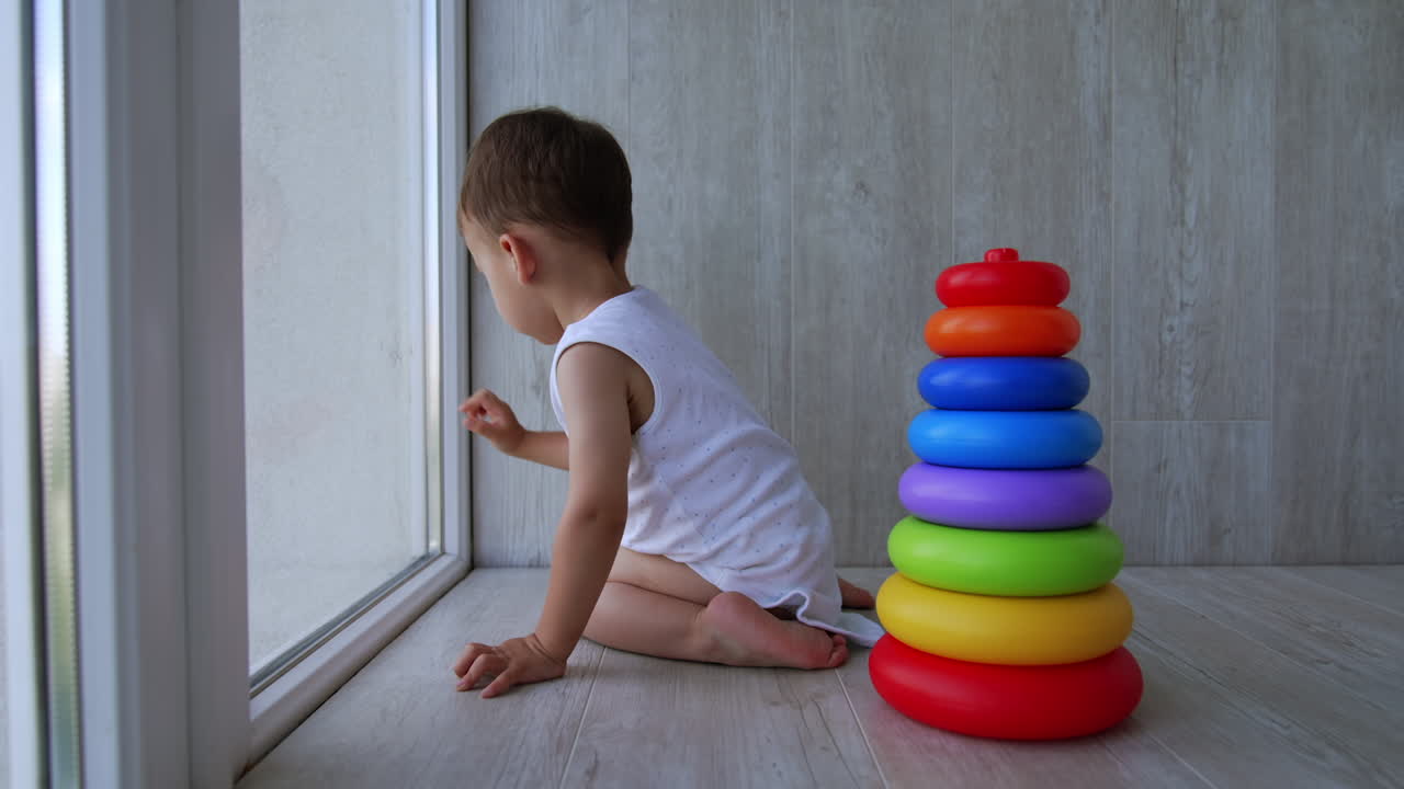 Lovely small kid in a white bodysuit sits at the window. Baby boy looks down attentively. Toy pyramid stands beside.