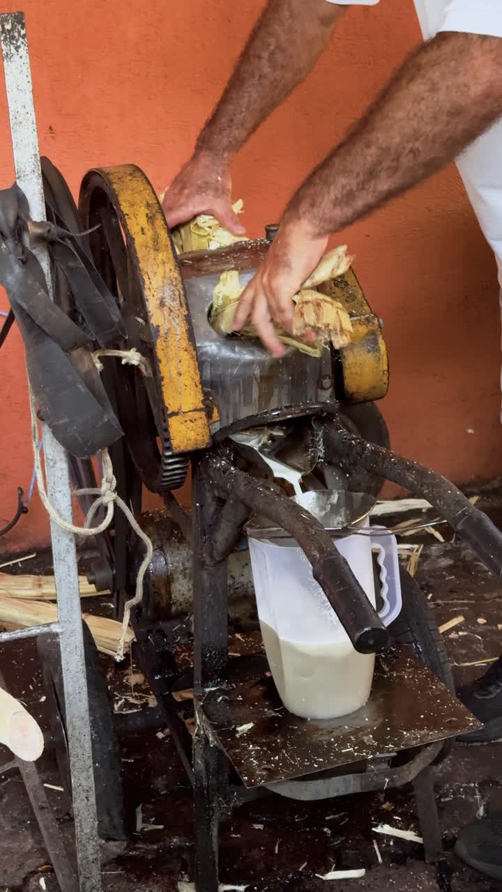 4K close-up showing a vendor’s arm operating an old, rusty sugarcane press in a crowded street market. The unsanitary yet functional machine creates juice while people gather around to buy.