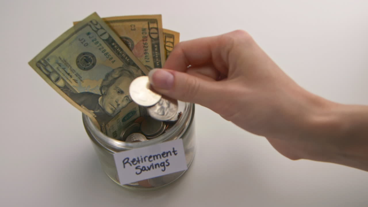 A caucasian woman's hand puts two American quarters into a &amp;quot;Retirement Savings&amp;quot; jar