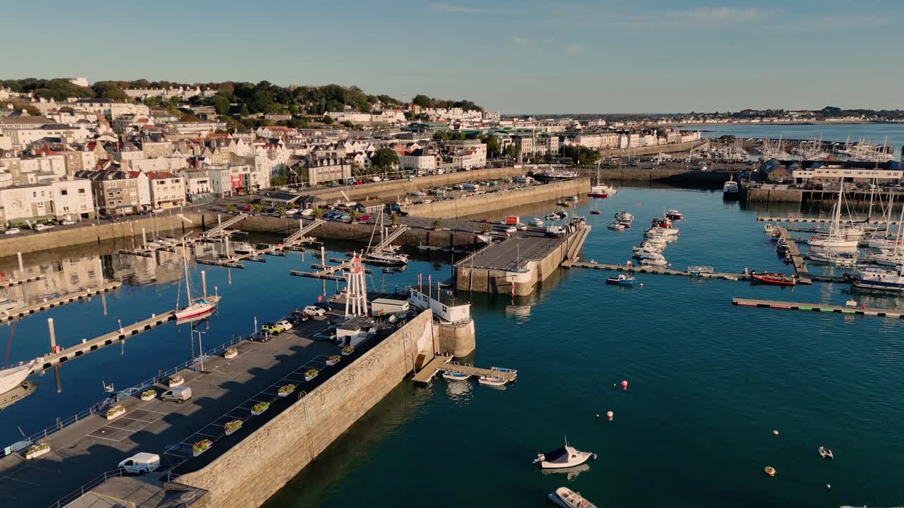 Drönarflygning över St Peter Port hamn, Guernsey, med Albert Marina, Victoria Marina och Crown Pier, samt båtar under kölhalning i sen eftermiddagssol, med staden och Belle Greve-bukten i fjärran som bakgrund.
