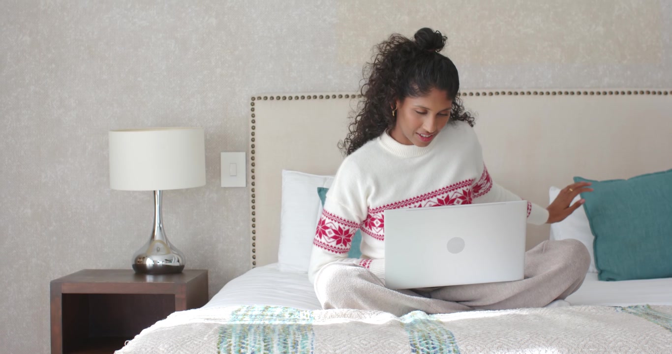 African American woman sitting on bed working on silver laptop, leaning closer for clearer view