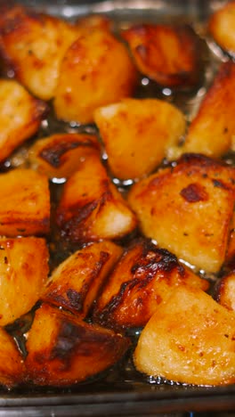 vertical video with camera moving along past golden roast potatoes in bubbling hot olive oil in glass dish. british cuisine traditionally served on sunday at lunch or dinner.