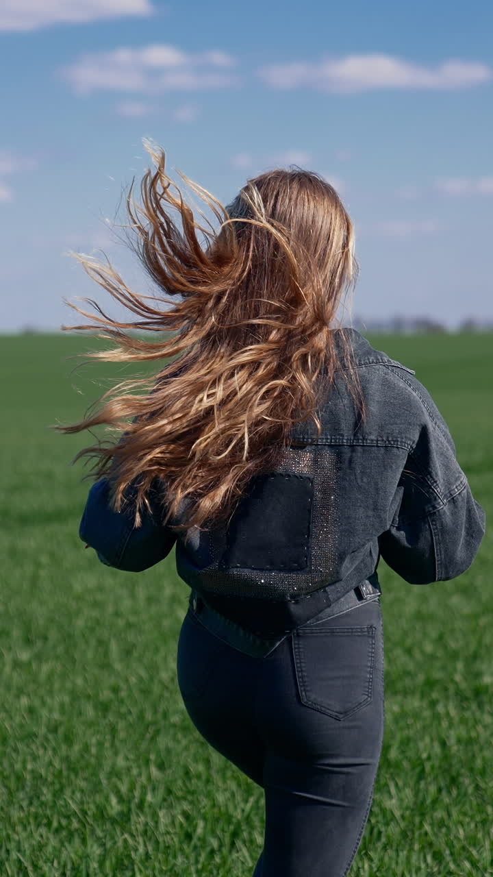 Girl is running on green field. Young woman with long fluttering hair in denim suit runs in nature and turning her cheerful face on camera. Rear view. Slow motion. Vertical video