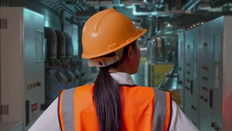 close up back view of a female engineer wearing safety helmet looking around while standing with arms akimbo in engine control room, work of electrical generators