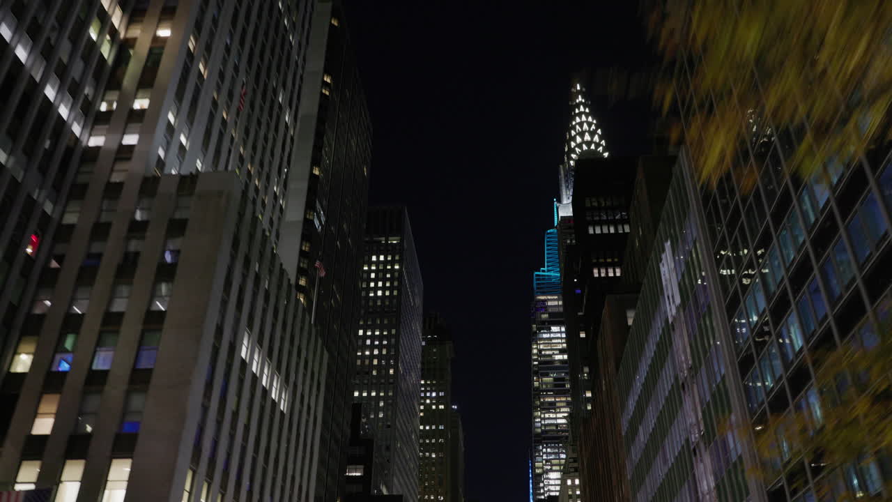 Passeio noturno pela rua de Manhattan com arranha-céus iluminados e o Edifício Chrysler à frente, perspectiva de movimento entre edifícios altos.