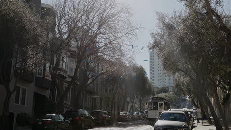 cable car moving uphill between parked cars and tall buildings in street scene