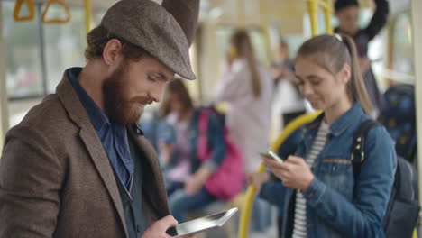 Young-Man-With-Beret-Using-Smartphone-Standing-In-The-Bus,-In-The-Background-A-Young-Girl-Standing-Chatting-With-Smartphone