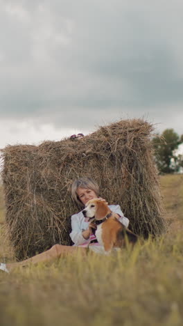 mujer de mediana edad con gafas de sol y traje casual se sienta contra la bala de heno en el campo, frotando suavemente la cabeza del perro, la hierba dorada, las colinas onduladas y el cielo nublado