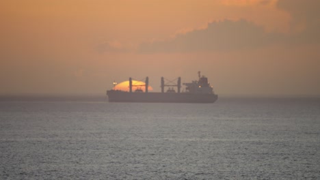 Weite-Kinoaufnahme-Eines-Frachtschiffs,-Das-Ladung-Transportiert-Und-In-Einen-Hafen-Einläuft,-Um-Dort-Zu-Entladen,-Während-Im-Hintergrund-Die-Sonne-Untergeht