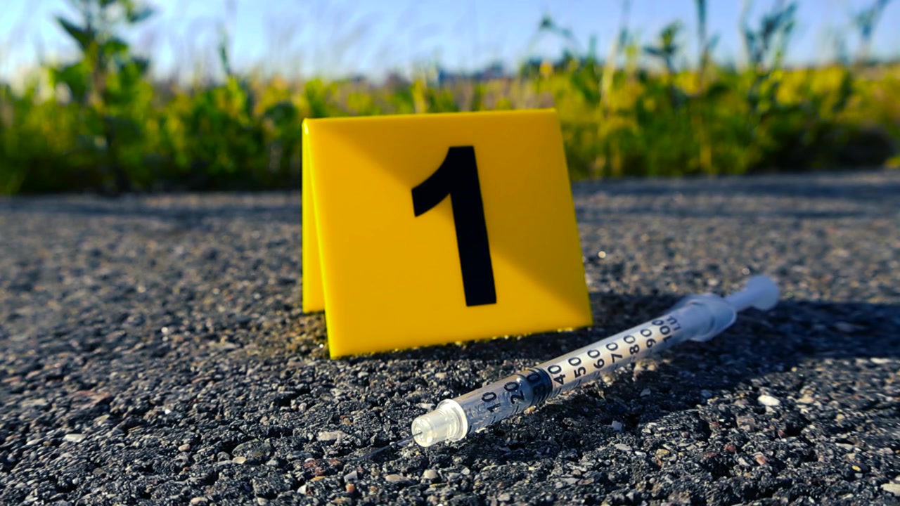 Primo piano di un segnalatore di prova giallo con il numero uno e una siringa usata su una strada asfaltata sulla scena di un crimine, che suggerisce un'indagine di polizia legata all'uso di droghe o a un omicidio