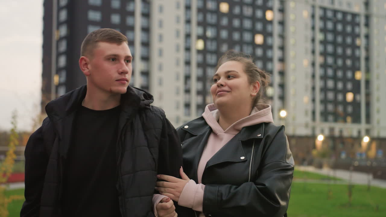 Boy looks unhappy as his sister gently holds his arm while smiling warmly during evening stroll near residential high-rise buildings with warm glowing lights through windows