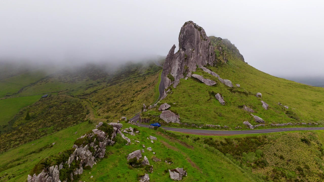 Auto che percorre una strada asfaltata passa vicino all'imponente formazione basaltica Rocha dos Frades sull'isola di Flores nelle Azzorre, Portogallo. Ripresa aerea con rotazione