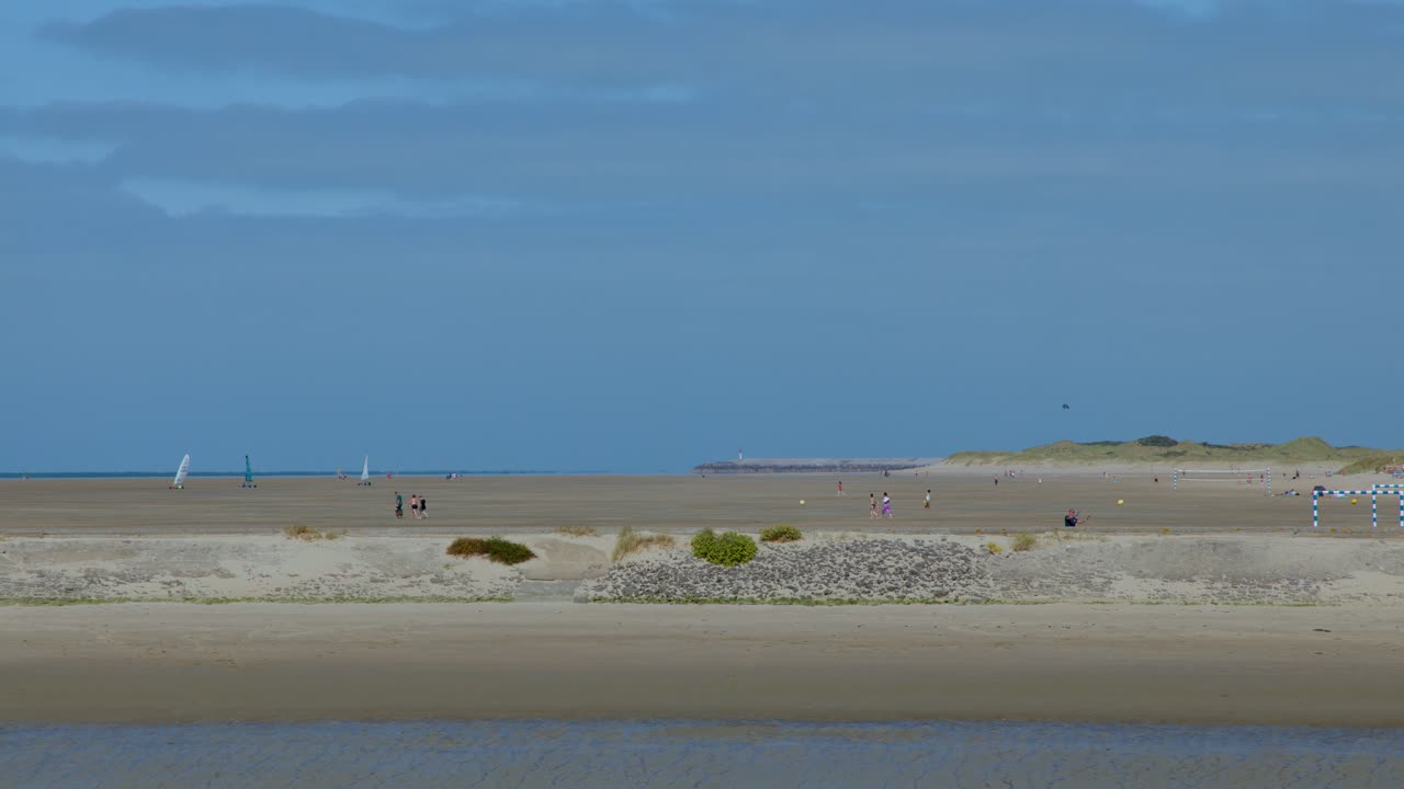 Wide static shot of sailboats moving across tranquil sandy beach under clear daylight sky