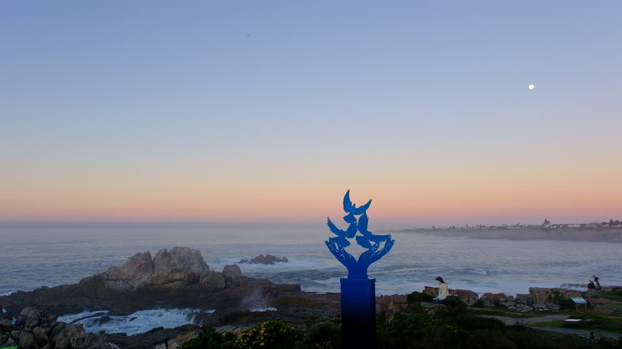 View of full moon setting during blue hour from Hermanus cliff path with statues