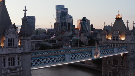 Low-flight-above-top-walkway-on-Tower-Bridge-in-evening.-Heading-to-group-of-skyscrapers-with-glass-facades.-London,-UK