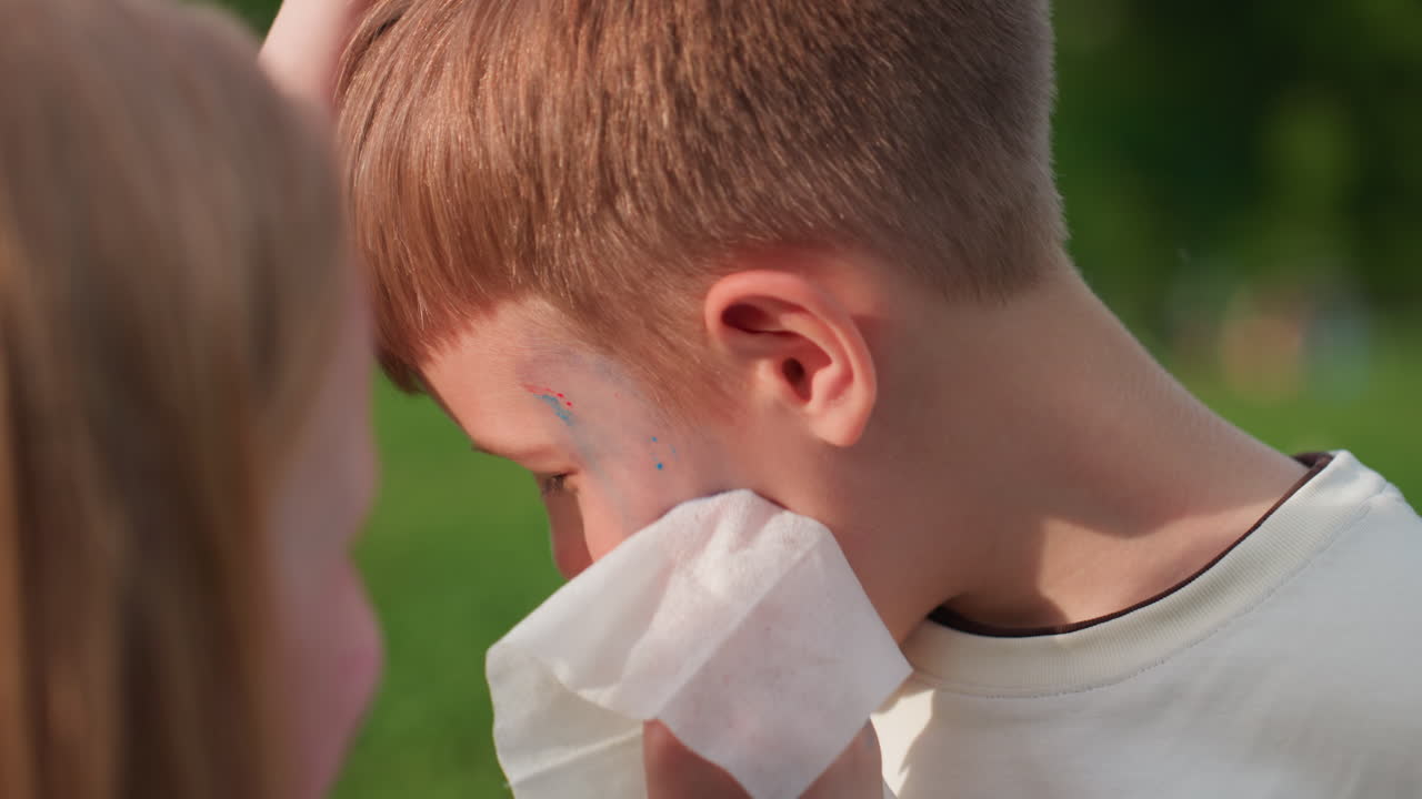 close up hands of girl cleaning paint from brother face with tissue in grassy park, warm light creating soft background blur, showing gentle sibling care and playful summer bonding