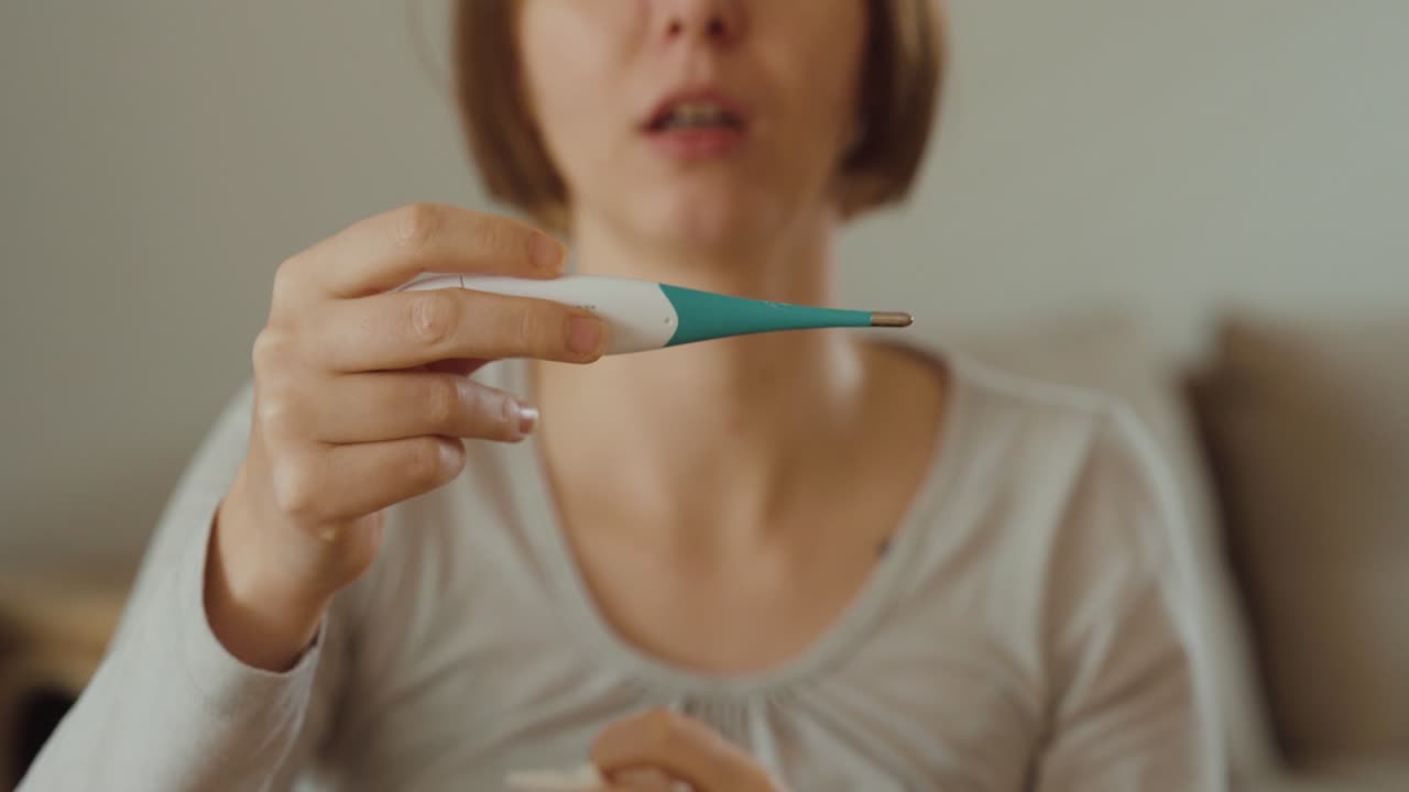 Eine Frau sitzt mit einem Thermometer zu Hause und putzt sich die Nase – Konzept für Erkältung, Grippesymptome, Krankheit und häusliche Genesung