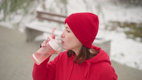 woman in red beanie and hoodie holding pink water bottle outdoors, preparing to drink, surrounded by snowy scenery and a blurred bench in serene winter setting