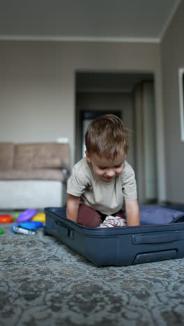 caucasian baby boy stands in front of a big suitcase opened in front of him on the floor. little kid gets into the suitcase. vertical video.