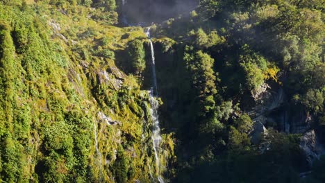 tiro inclinado hacia abajo de una cascada épica entre paredes verdes de montaña iluminadas por el sol - parque nacional fiordland, nueva zelanda