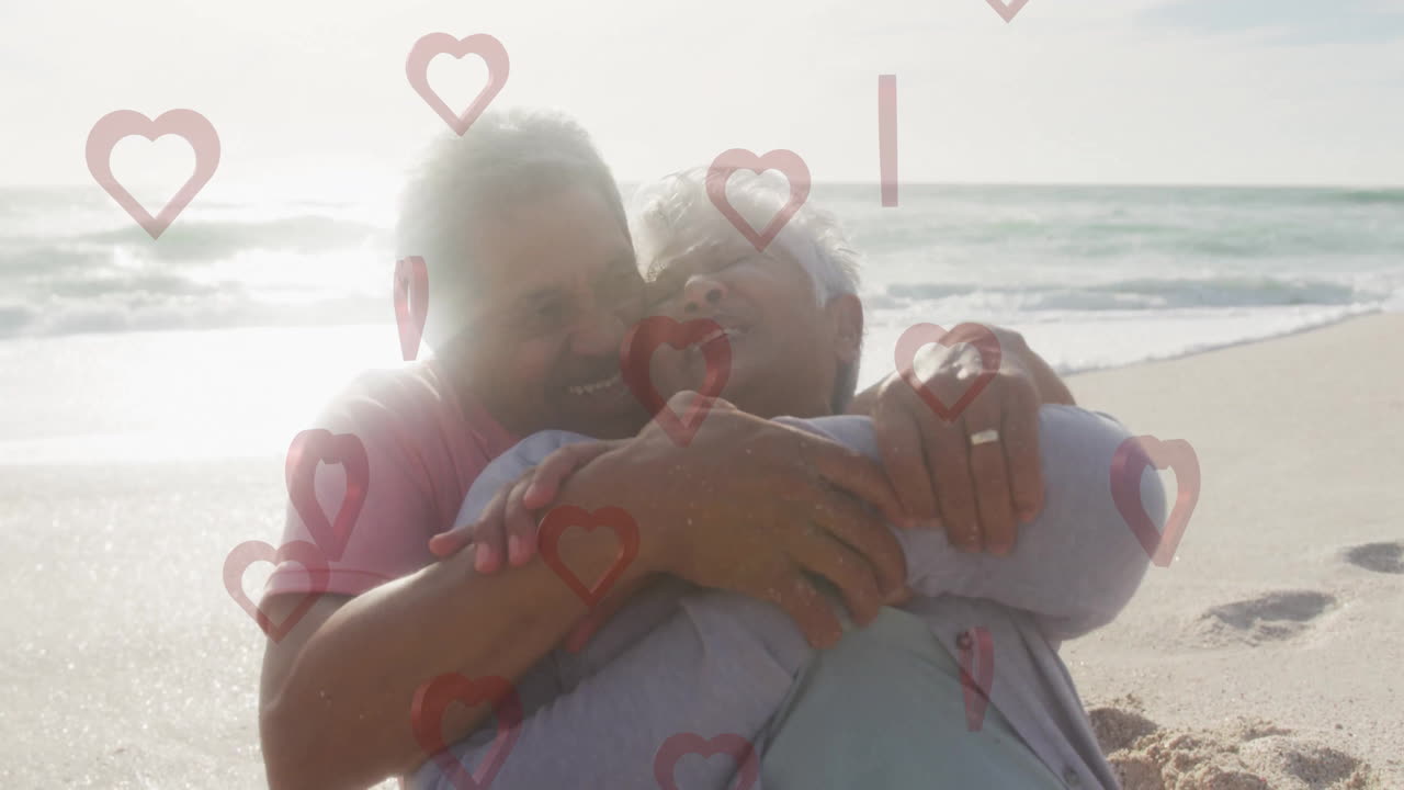 Embracing on beach, elderly couple with heart animation overlay in background