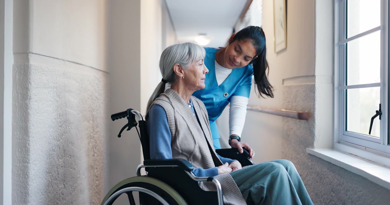 Caregiver Talking to Elderly Woman in Wheelchair in Hospital Hallway