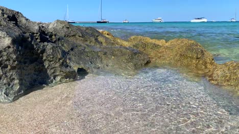 Summer-dream-vacation-destination-in-Formentera-island-Ses-Illetes-beach-Ibiza-Spain,-transparent-turquoise-water-and-luxury-yachts-on-holiday,-waves-hitting-rocks,-4K-shot