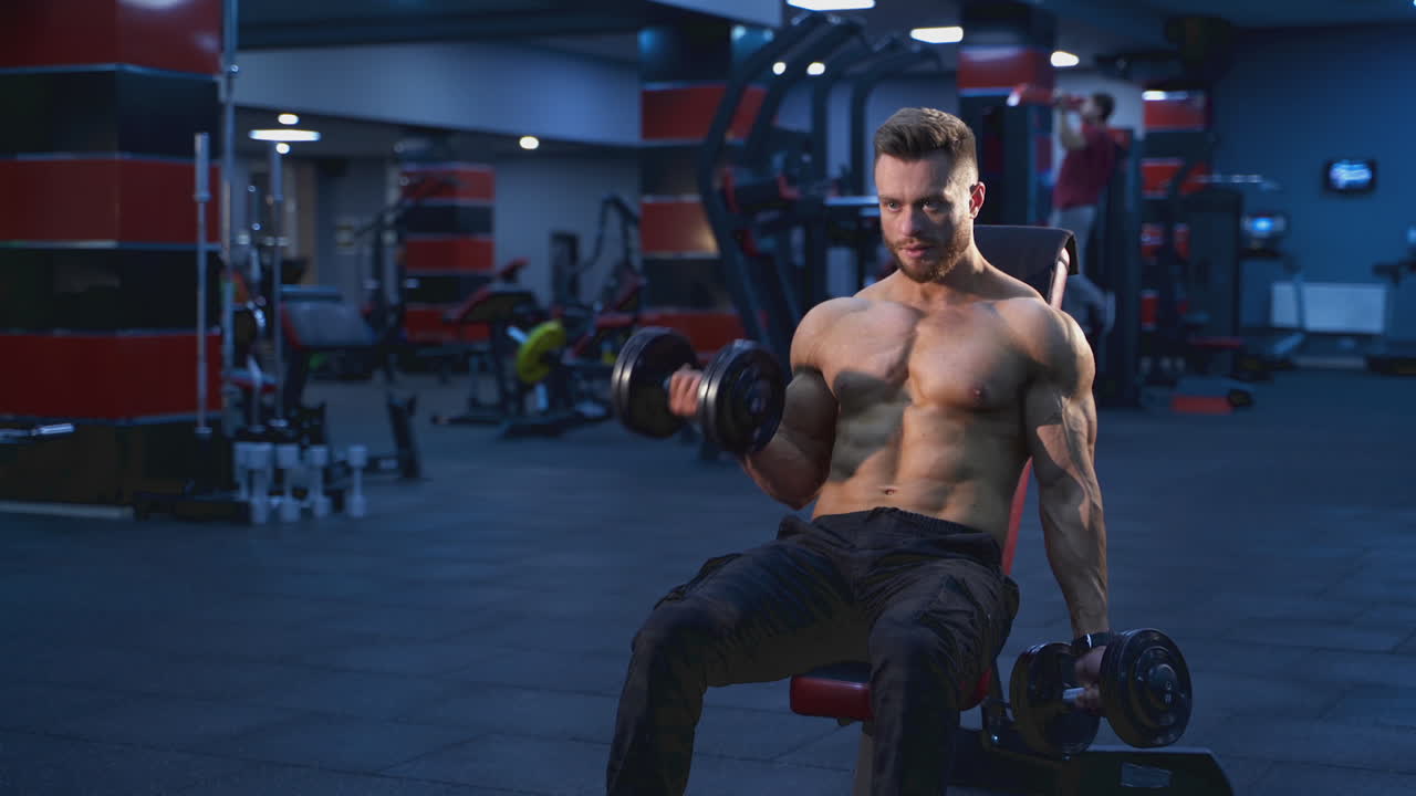 Bodybuilder che si allena in una palestra moderna. L'atleta è seduto su una sedia e pompa i muscoli delle braccia con i manubri in un centro sportivo. Allenamento quotidiano