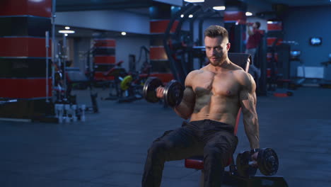 bodybuilder training in the modern gym. sportsman is sitting on a chair and pumping up arm muscles with dumbbells in sports centre. daily workout.